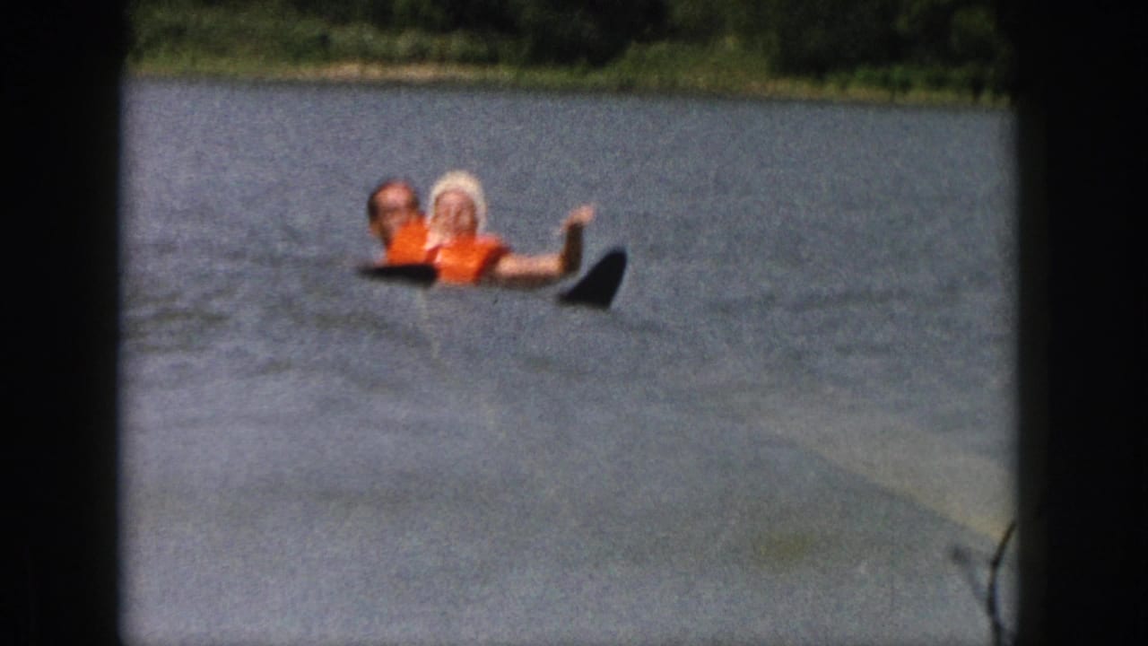 1961 Michigan home-movie frame showing two people in orange life vests floating on a lake before a water-ski attempt.