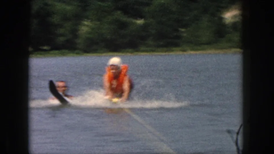 A water-skier in an orange life vest and white hat is pulled across a Michigan lake as a helper or swimmer appears nearby.