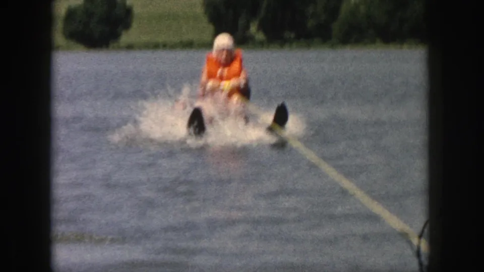 The woman in an orange life vest and white hat appears to balance low on water skis while being pulled across the lake.