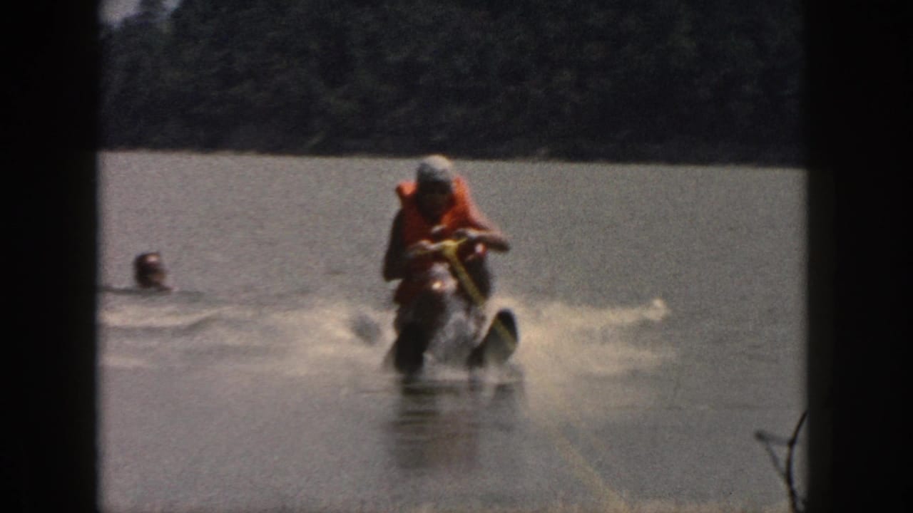 1961 Michigan 8mm frame showing a water skier in an orange life vest moving across a wooded lake with spray behind him.