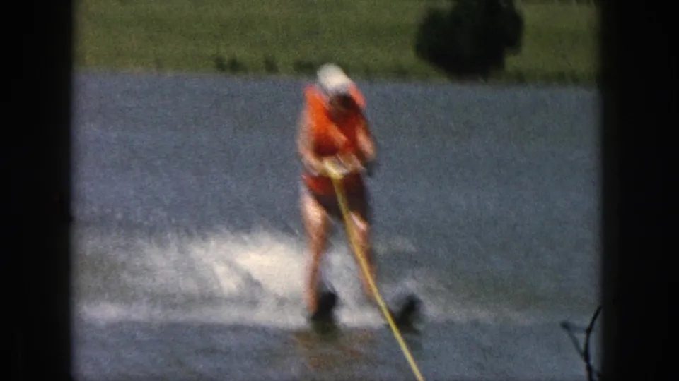 1961 Michigan home-movie frame showing a water skier leaning into the tow rope, with lake water and shoreline behind him.