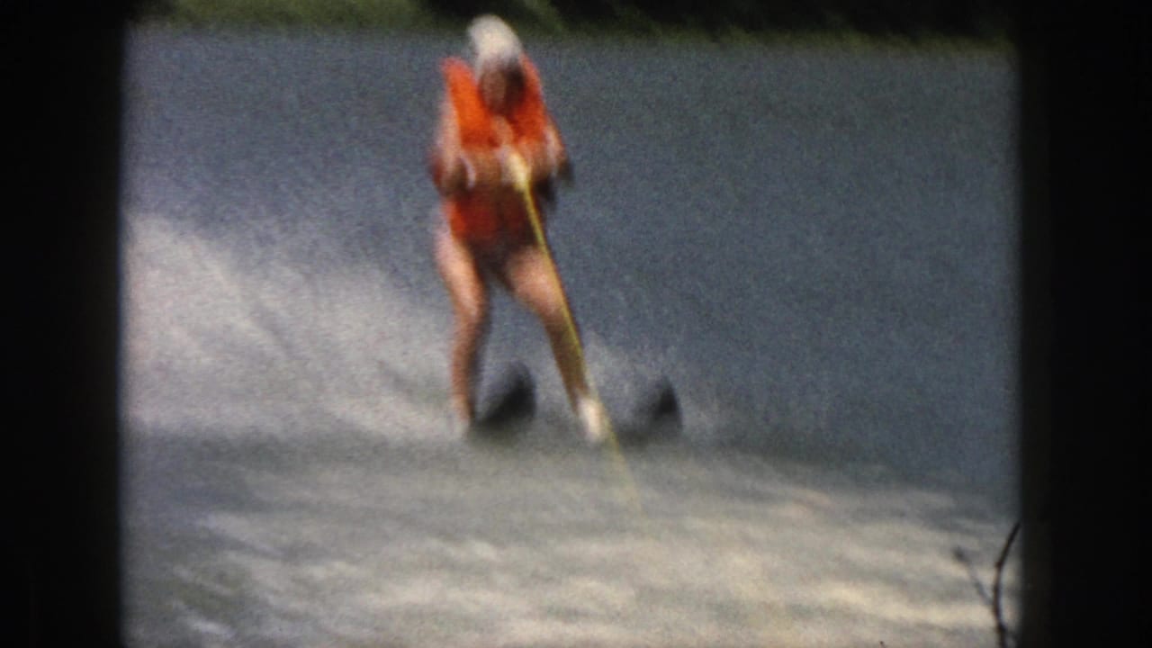In Michigan 1961 home-movie footage, a woman in an orange life vest and white swim cap skis across a lake behind a yellow rope.