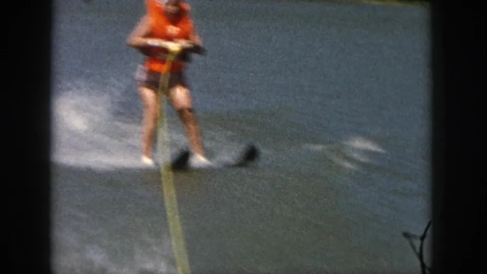 A woman in an orange life vest holds a yellow tow rope while water skiing on a Michigan lake in 1961 home-movie footage.