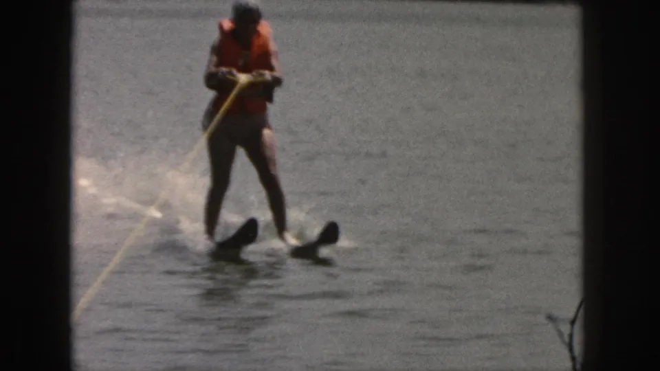 A woman in an orange life vest stands upright on water skis, holding a yellow tow rope on a Michigan lake in 1961 footage.