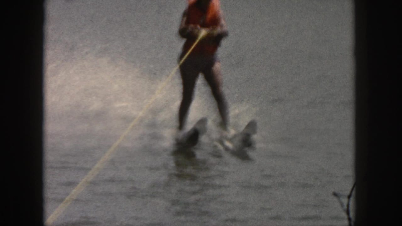 1961 Michigan home-movie frame showing a water skier in an orange life vest and dark swimwear behind a tow rope on a lake.