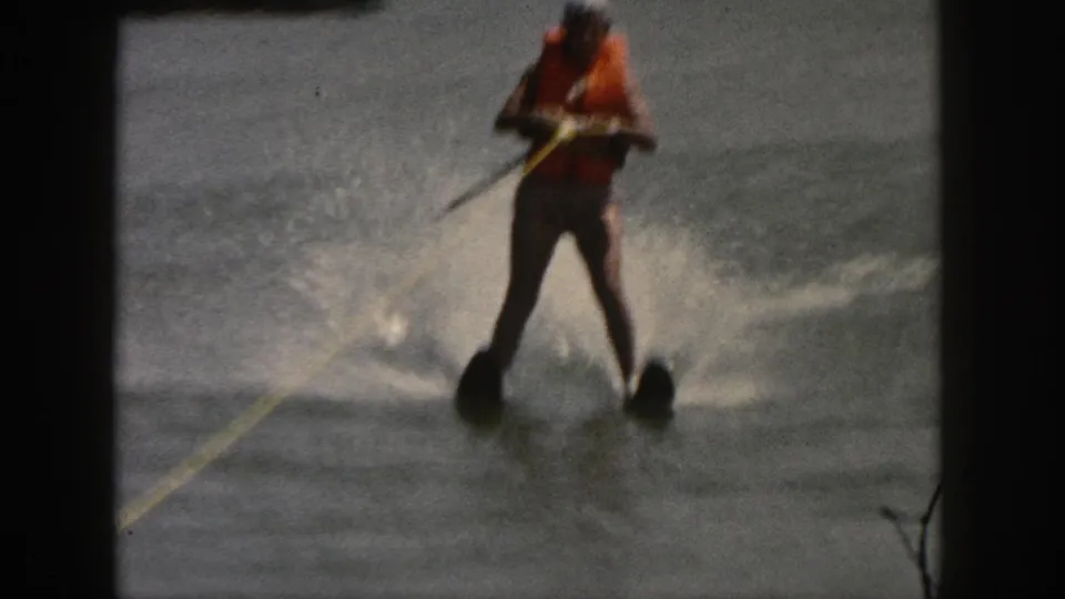 Grainy 8mm frame of a 1961 Michigan water skier facing the camera, with an orange life vest, yellow tow rope, and splashing wake.