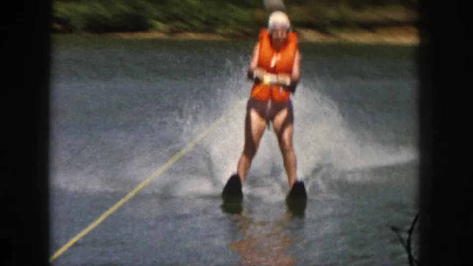 1961 Michigan 8mm still of a skier riding upright in an orange life vest, with river spray and a wooded shoreline in the background.
