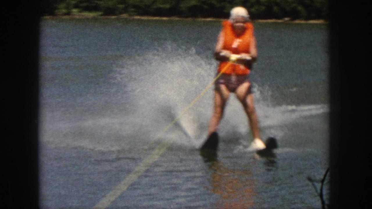 1961 Michigan home-movie frame showing a water skier in an orange life vest gliding on twin skis across calm lake water.