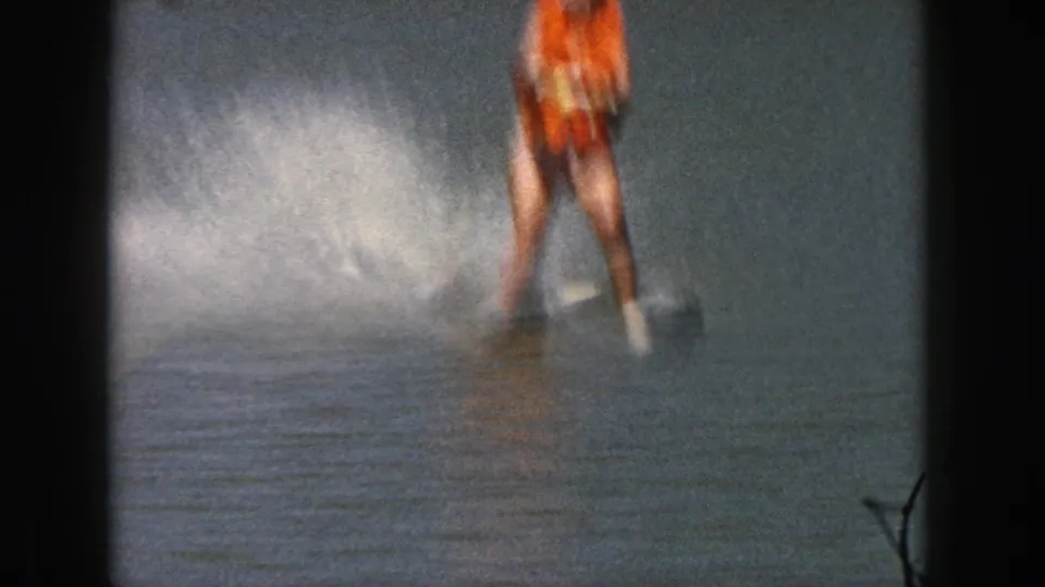 Cropped 1961 Michigan home-movie frame showing the skier’s lower body, orange vest, skis, and a large burst of spray.