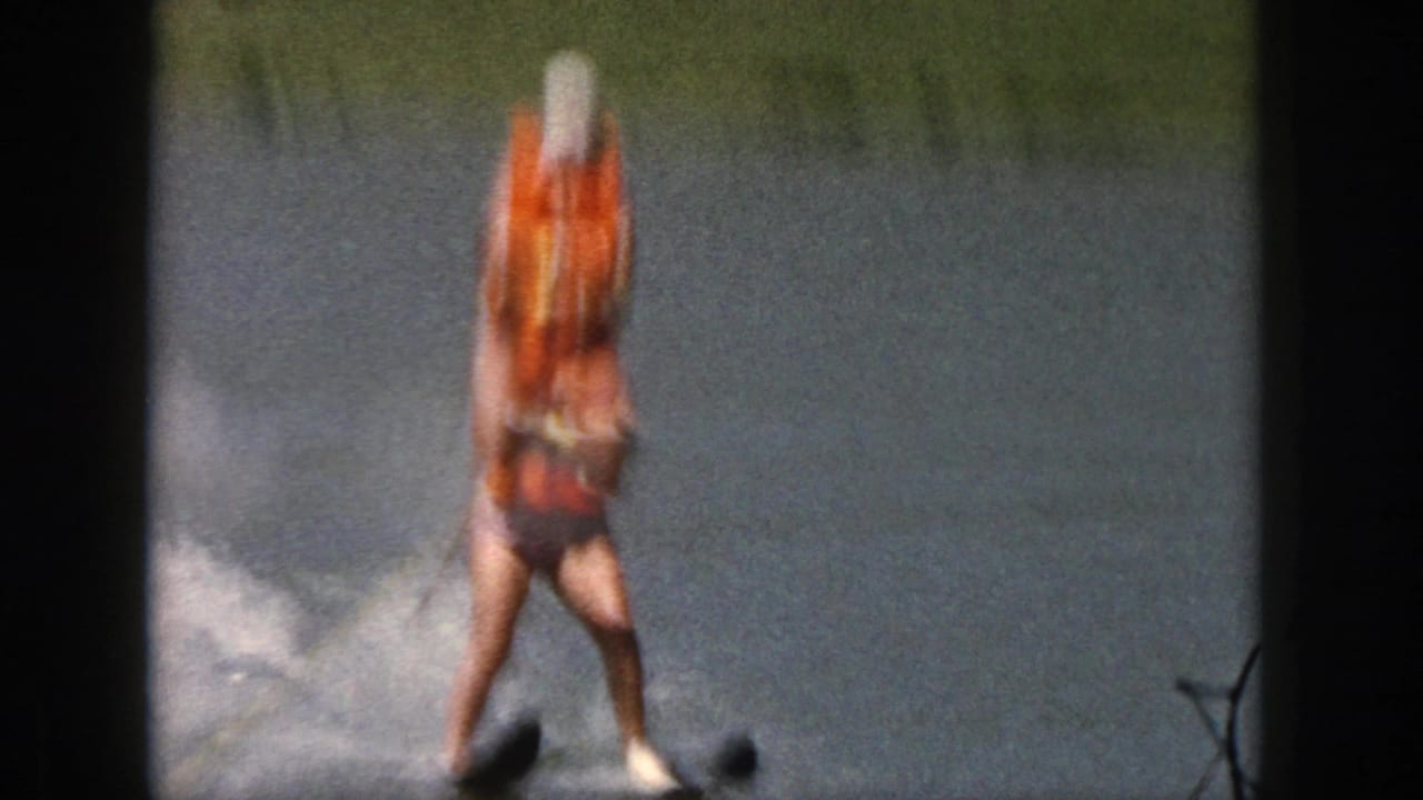 1961 Michigan 8mm frame of a water skier in an orange life vest and white cap gliding across a lake.