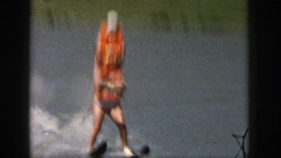1961 Michigan 8mm frame of a water skier in an orange life vest and white cap gliding across a lake.