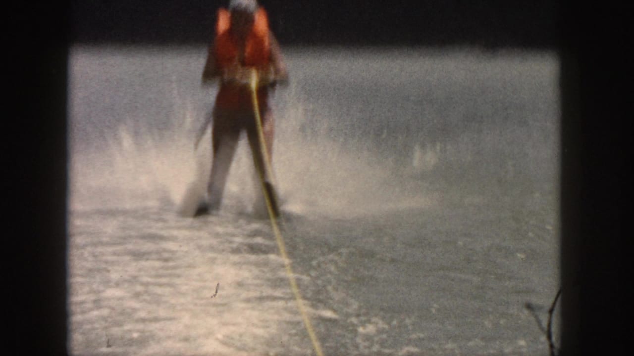 1961 Michigan 8mm frame of a water skier in an orange life jacket and swim cap gliding on a calm river.