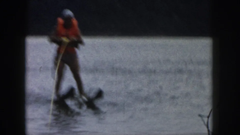 1961 Michigan home-movie frame of the skier leaning low as the skis and wake disturbance appear in front.