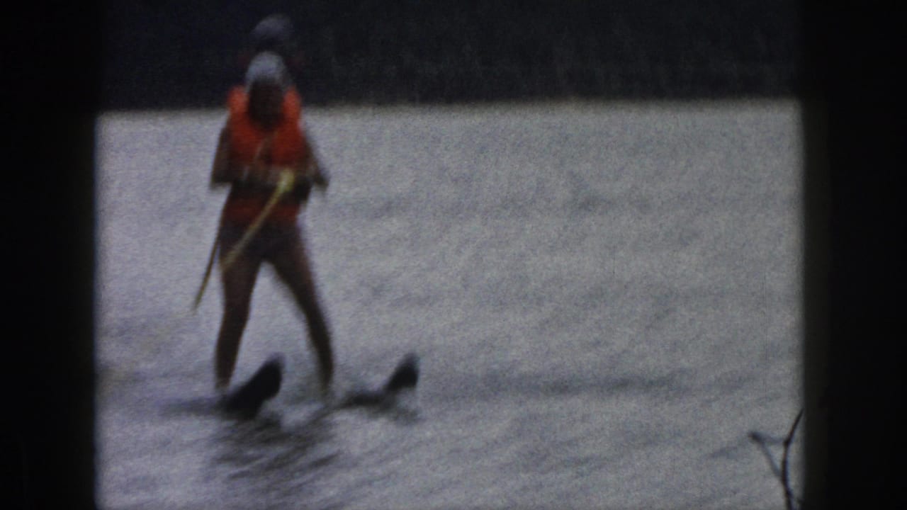1961 Michigan home-movie frame with a water skier facing the camera, holding a rope while moving over the lake.