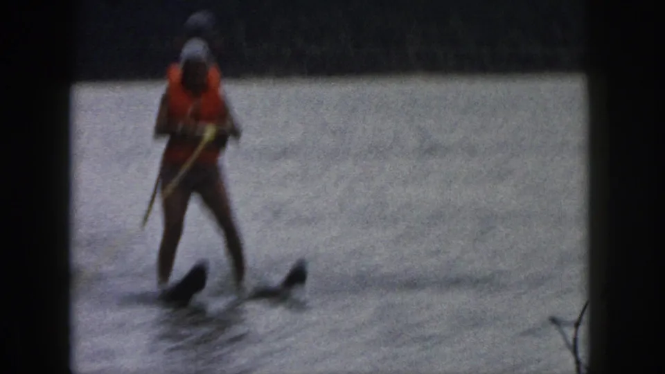 1961 Michigan home-movie frame with a water skier facing the camera, holding a rope while moving over the lake.