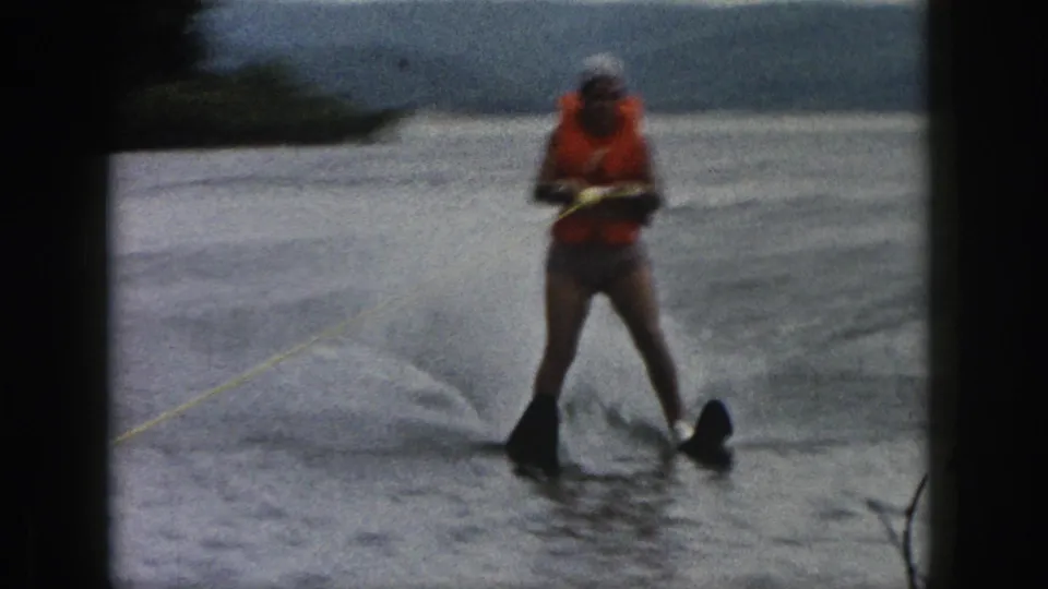 1961 Michigan home-movie frame with a skier in an orange vest holding a tow rope as spray trails behind.
