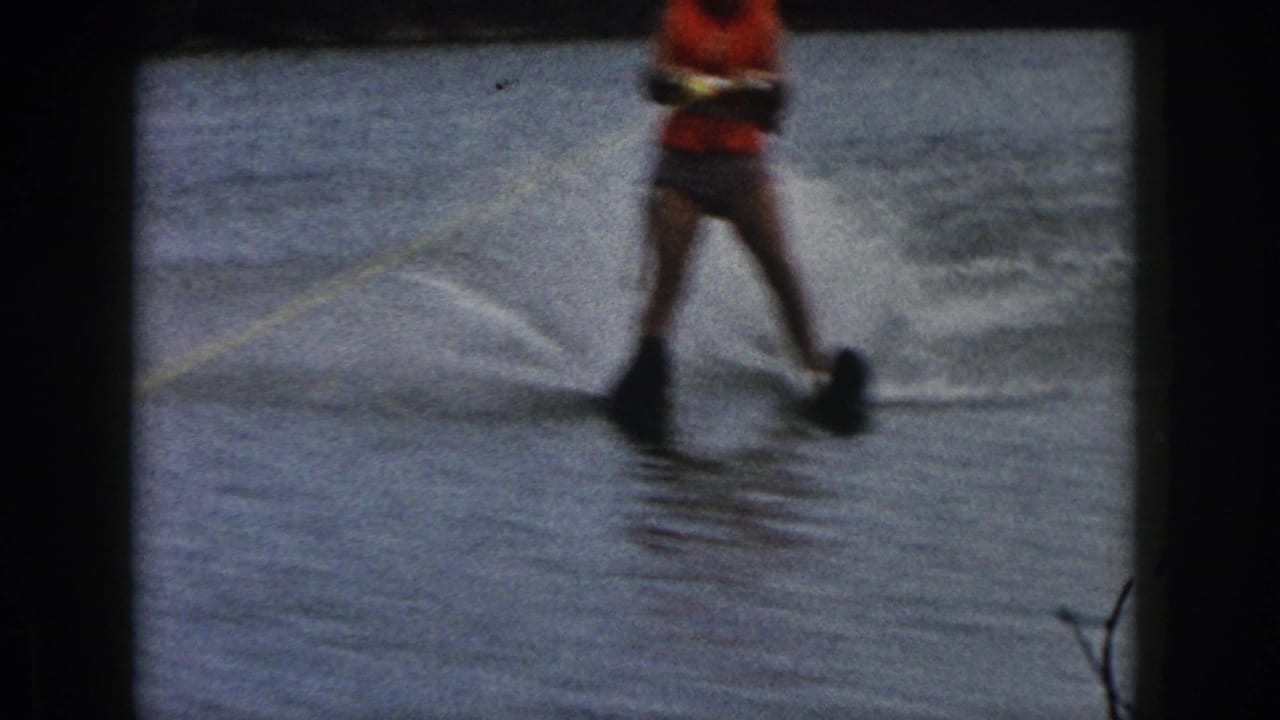1961 Michigan home-movie frame showing a woman water skiing on a lake, partially cropped, with black skis and a yellow tow rope.