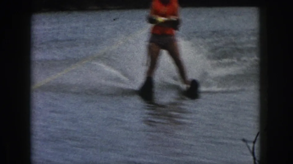 1961 Michigan home-movie frame showing a woman water skiing on a lake, partially cropped, with black skis and a yellow tow rope.