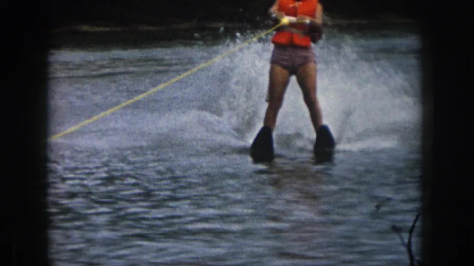 1961 Michigan frame of a woman on black water skis holding a yellow tow rope, with lake spray rising behind her.