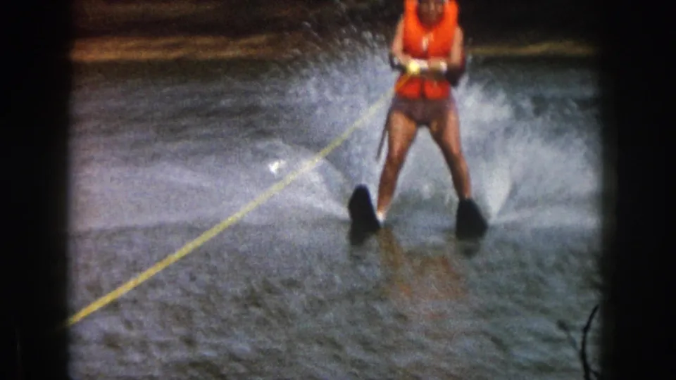 1961 Michigan home-movie frame of a cropped woman water skier in an orange life preserver, pulled by a yellow rope across the lake.