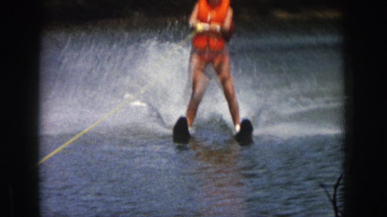 1961 Michigan home-movie frame showing a water skier in an orange life vest creating spray while holding a yellow tow rope.