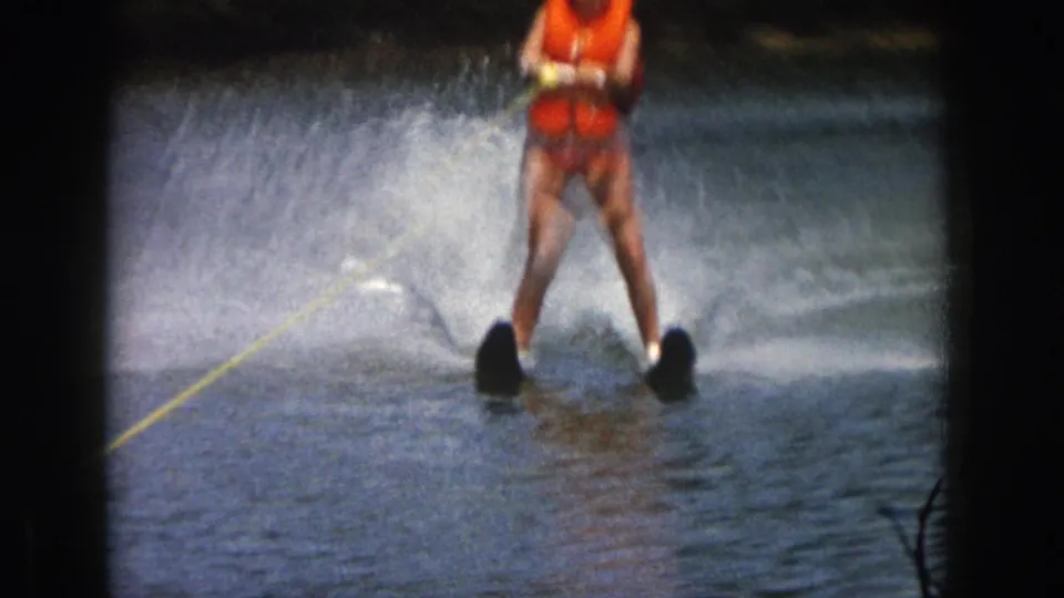 1961 Michigan home-movie frame showing a water skier in an orange life vest creating spray while holding a yellow tow rope.