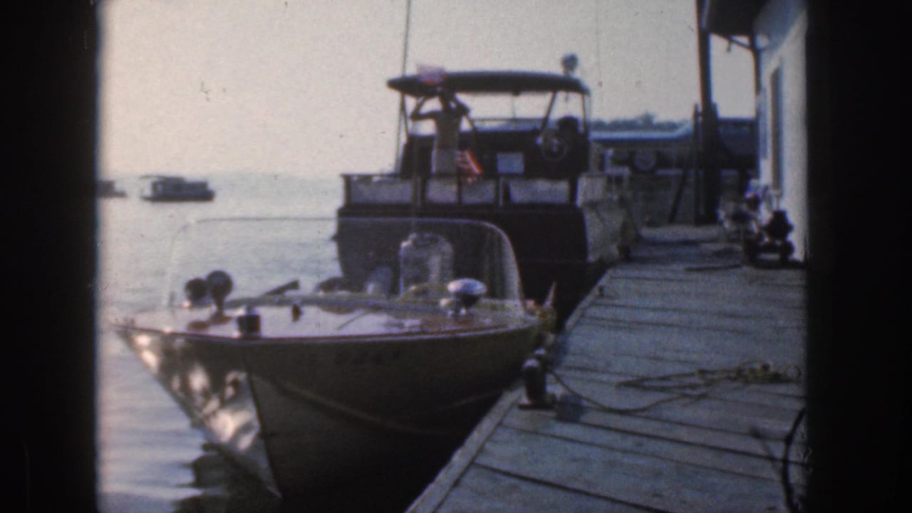 1961 Michigan home-movie frame showing a small windshield motorboat tied at a dock, with a larger cruiser and calm water behind it.