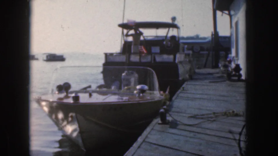 1961 Michigan home-movie frame showing a small windshield motorboat tied at a dock, with a larger cruiser and calm water behind it.