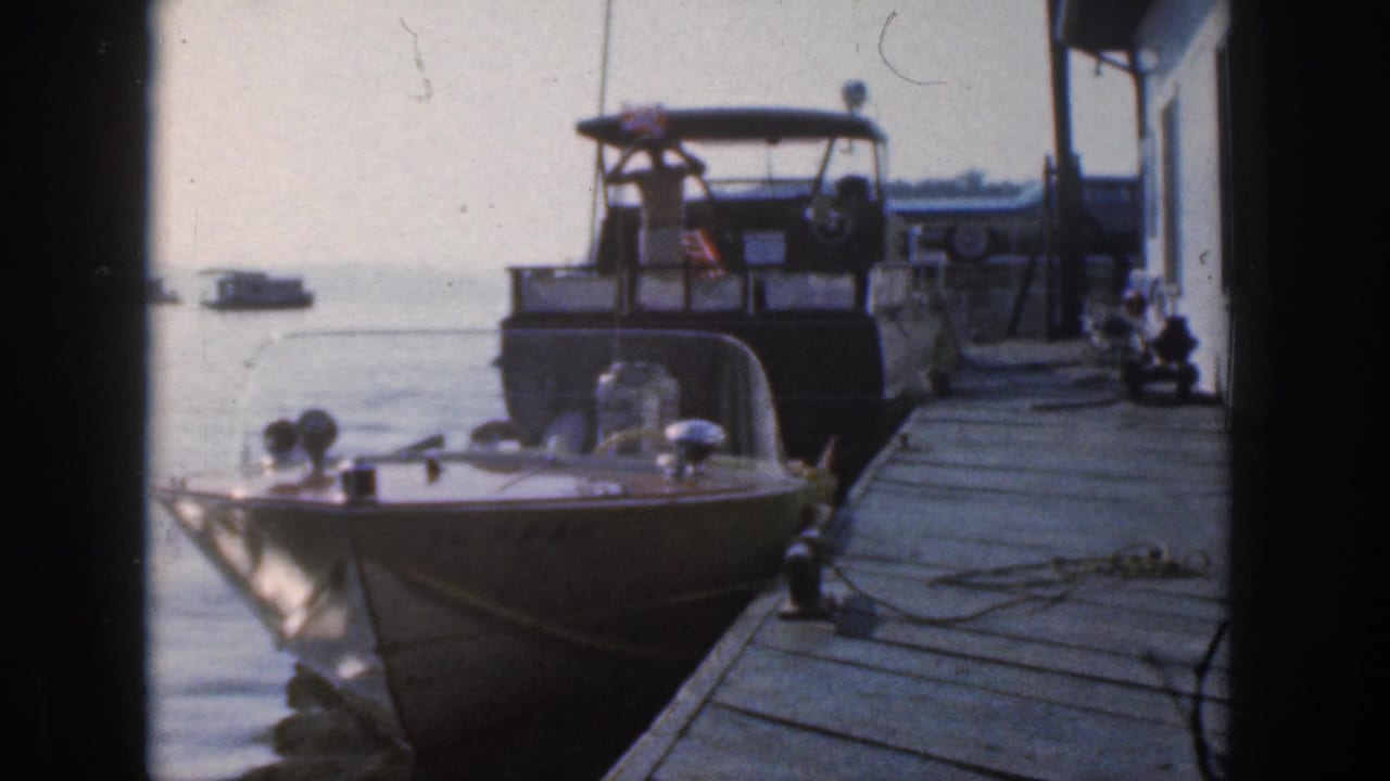 Foreground speedboat and dock planks with a larger boat and flag in the background.