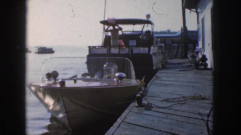 A dockside 8mm view of a foreground speedboat, wooden planks, mooring lines, and a larger cruiser flying an American flag.