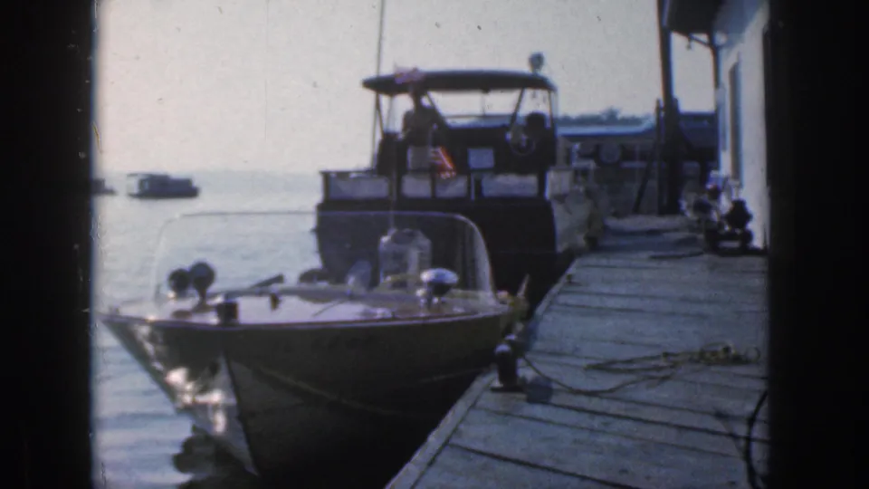 1961 Michigan 8mm footage frame with two moored motorboats, dock planks, rope, water, and a distant boat on the lake.