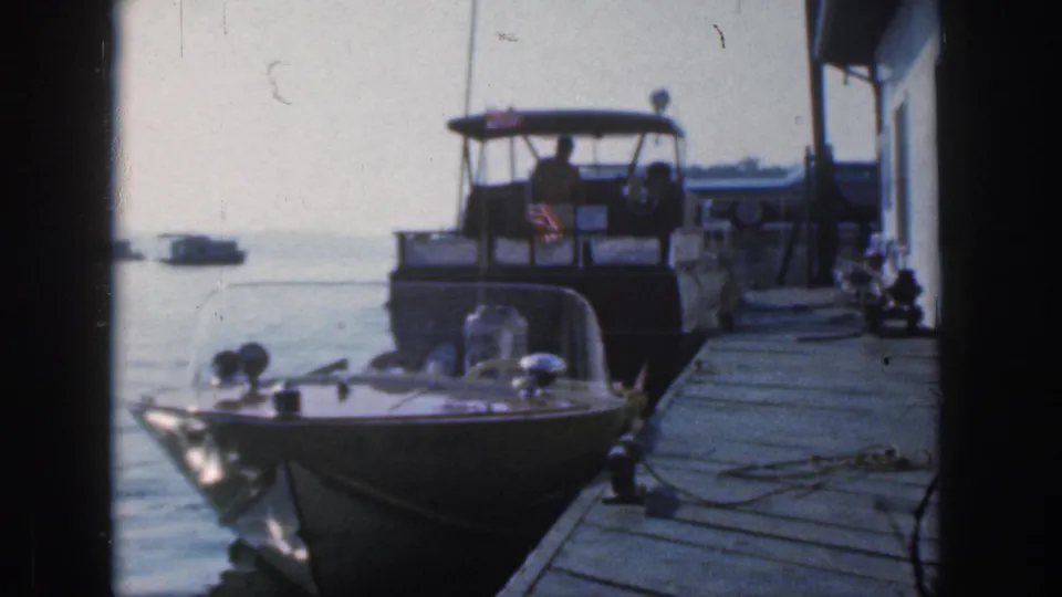 Home-movie frame from Michigan in 1961 showing a small docked speedboat, a larger cruiser, mooring ropes, and still lake water.
