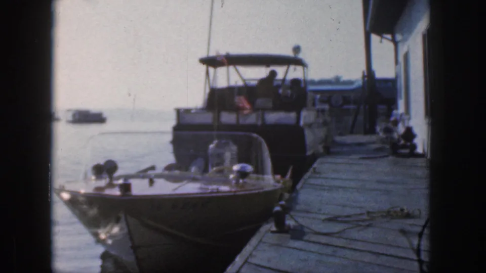 1961 Michigan 8mm frame showing a speedboat in the foreground and a cabin cruiser tied to a wooden pier on calm water.