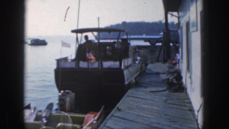 Home-movie frame from Michigan in 1961 showing a docked cabin cruiser, wooden pier, American flag, and distant boat on calm water.