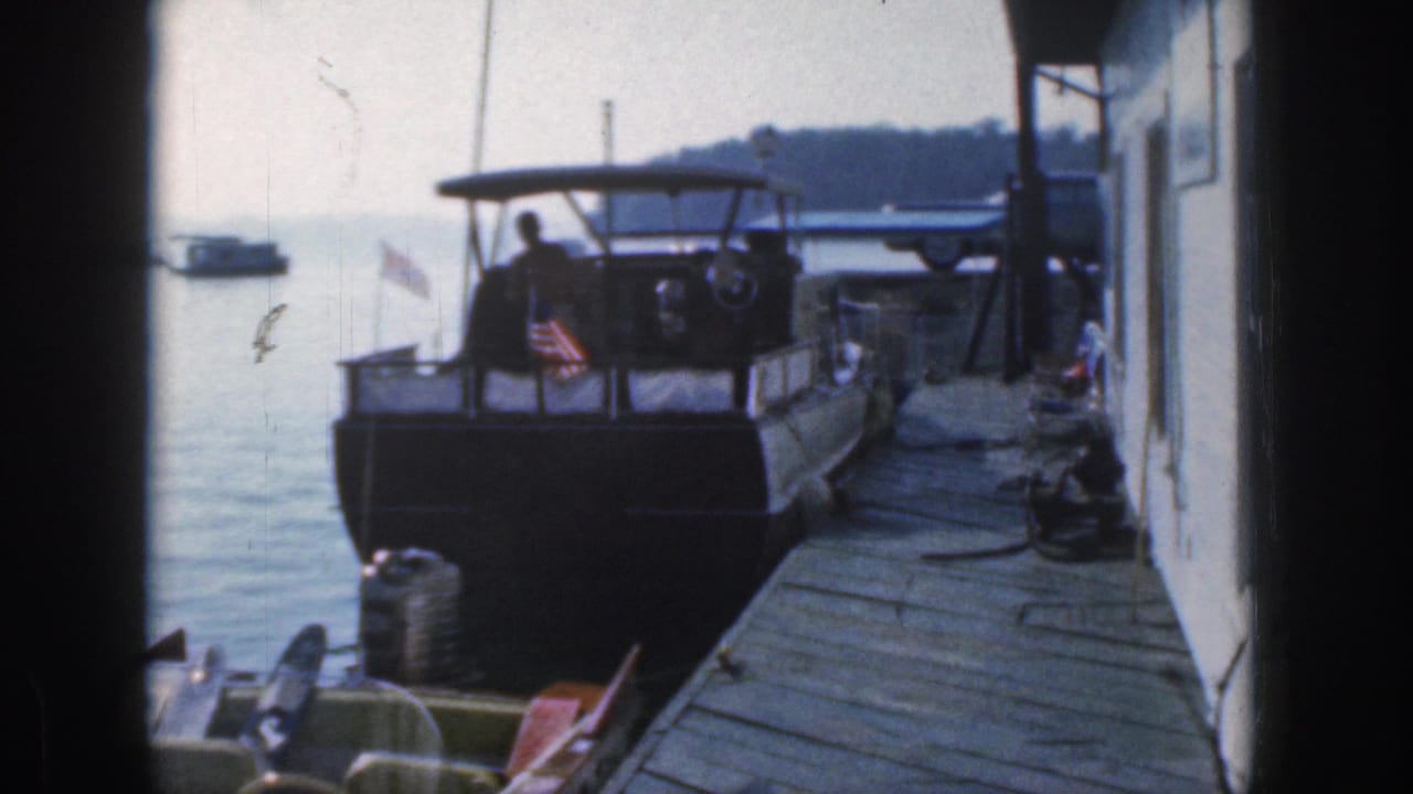 1961 Michigan 8mm frame showing a man on a docked canopy motorboat near a wooden pier, with a small flag and lake water behind.