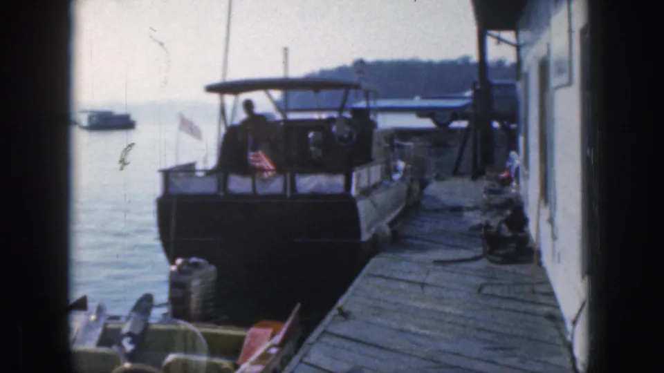 1961 Michigan 8mm frame showing a man on a docked canopy motorboat near a wooden pier, with a small flag and lake water behind.