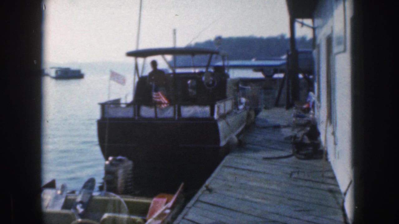 A 1961 Michigan home-movie frame shows a man silhouetted on a docked motorboat beside a wooden pier, with lake water and shoreline beyond.
