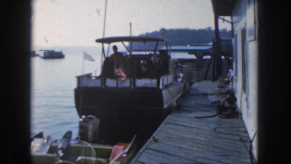 The 8mm frame shows the same Michigan dock scene with a canopy motorboat, small flag, wooden planks, and a distant boat on the lake.