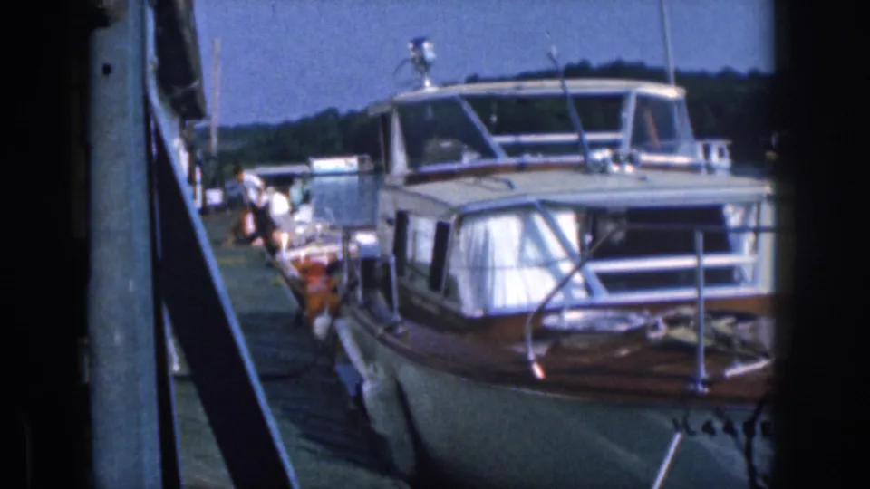 A 1961 Michigan 8mm frame shows a larger cabin cruiser tied along the dock, with railings, windows, shoreline, and waterfront structures visible.
