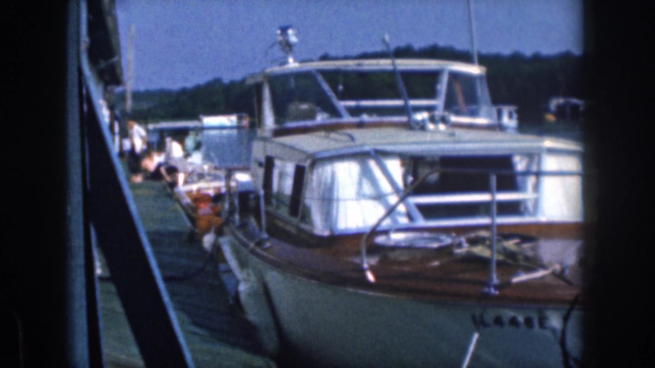 1961 Michigan 8mm frame showing a classic cabin motorboat tied at a lakeside dock, with people faintly visible behind it.