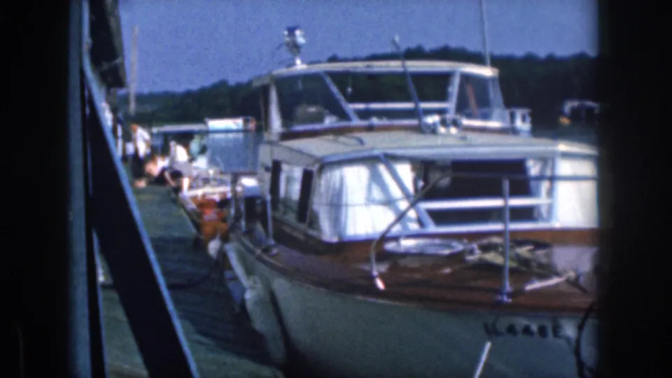 1961 Michigan 8mm frame showing a classic cabin motorboat tied at a lakeside dock, with people faintly visible behind it.