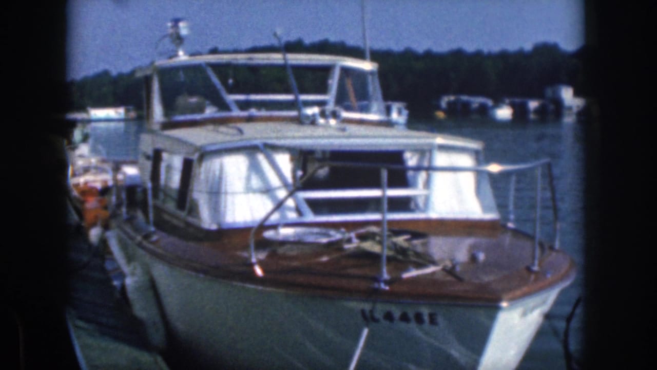 1961 Michigan home-movie frame of a moored white motorboat with wood trim, chrome rails, curtains, and distant boats on the water.