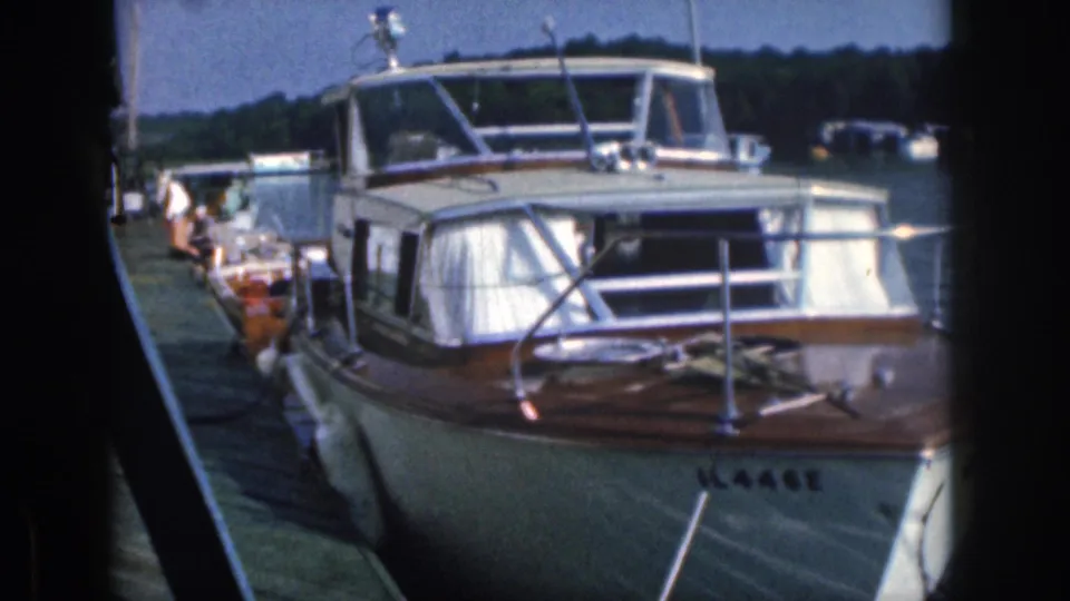 1961 Michigan 8mm frame showing the docked motorboat from the bow, with people along the dock and other boats across the lake.