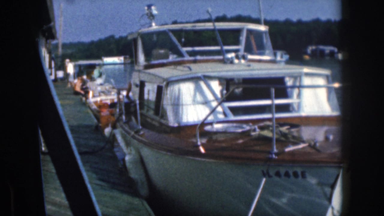 1961 Michigan home-movie frame showing a white wood-trimmed motorboat at a dock with people and other boats in the background.