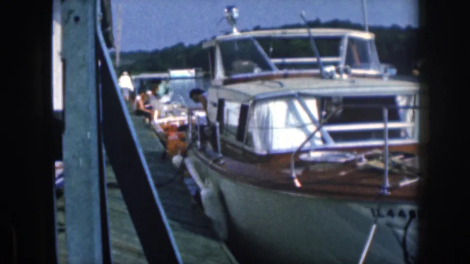 1961 Michigan 8mm frame of a motorboat moored close to a wooden pier, with people and small boats visible beyond it.
