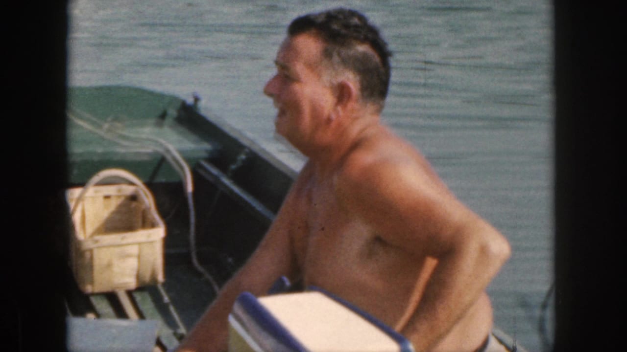 In 1961 Michigan home-movie footage, a shirtless adult man sits side-on in a small green motorboat with calm water and a woven basket visible.