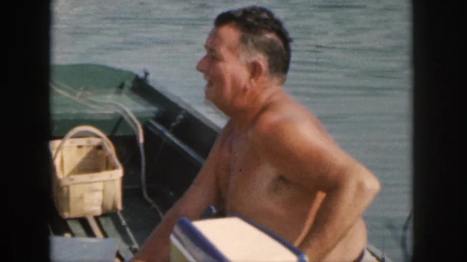 In 1961 Michigan home-movie footage, a shirtless adult man sits side-on in a small green motorboat with calm water and a woven basket visible.