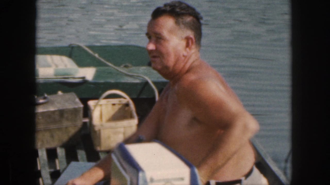 Shirtless man steering a small green boat with a white cooler in the foreground.
