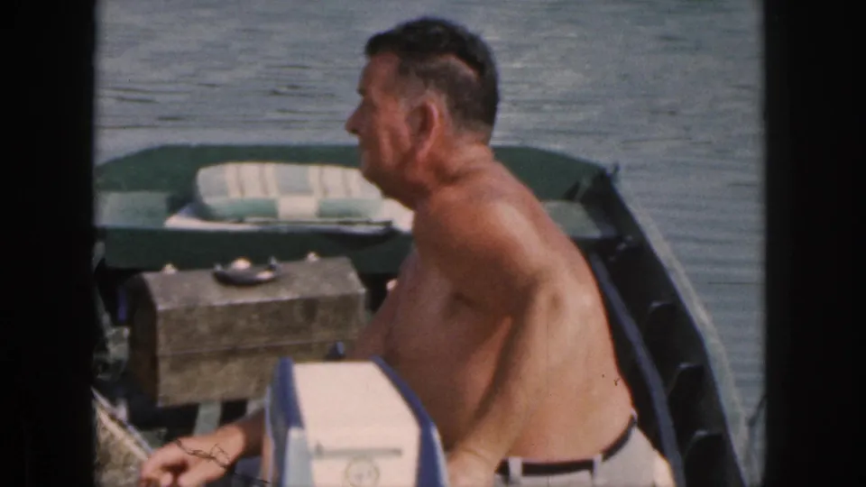 The 8mm frame shows the man in side profile inside the small green boat, with calm water, a wooden box, and onboard gear visible.