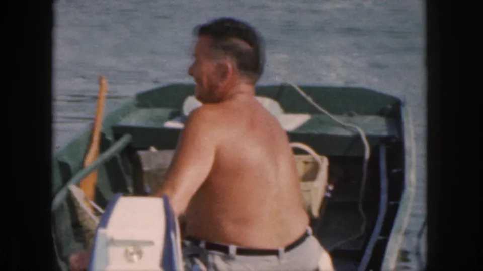 A rear-side view shows the shirtless man in the small green boat, with an oar, woven basket, and calm water in the 1961 Michigan scene.